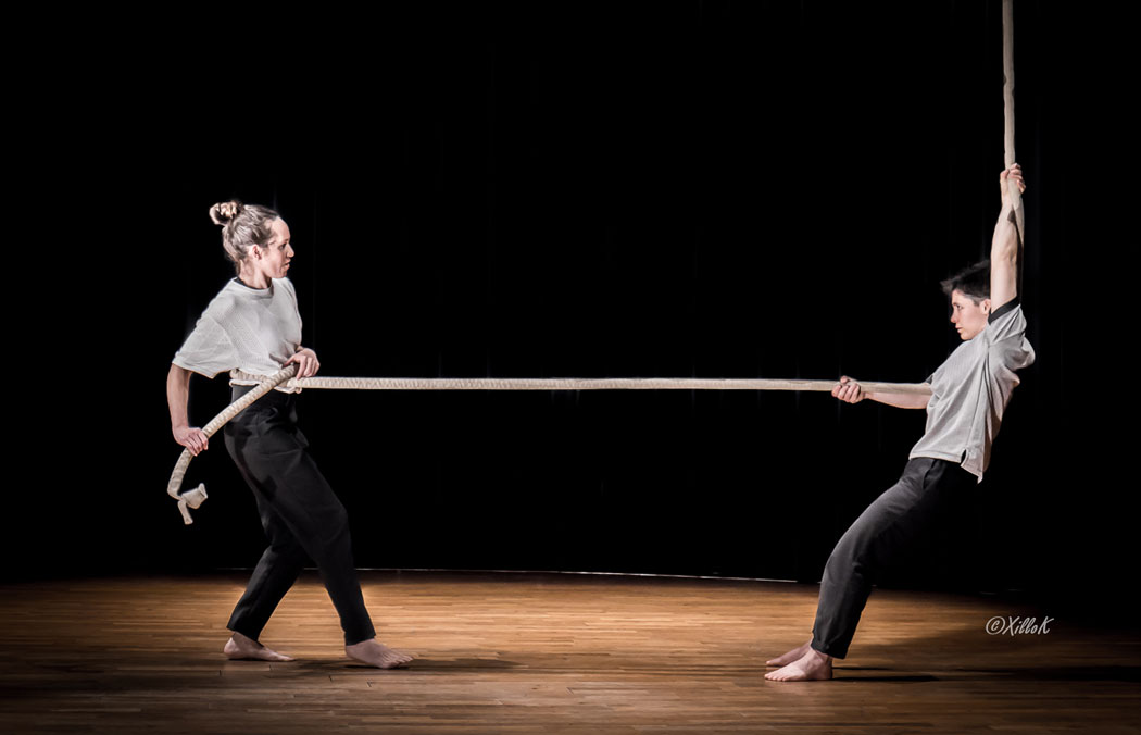 Crédit Karine Crozes. Photo du spectacle. En salle, sur scène.
2 circassiennes sont de chaque côtés d'une corde épaisse et la tire en faisant jouer leur contrepoids.
La corde et leurs regards symbolisent les tensions dans leur relation.
La lumière est présente en une douche de couleur chaude en fond de scène.