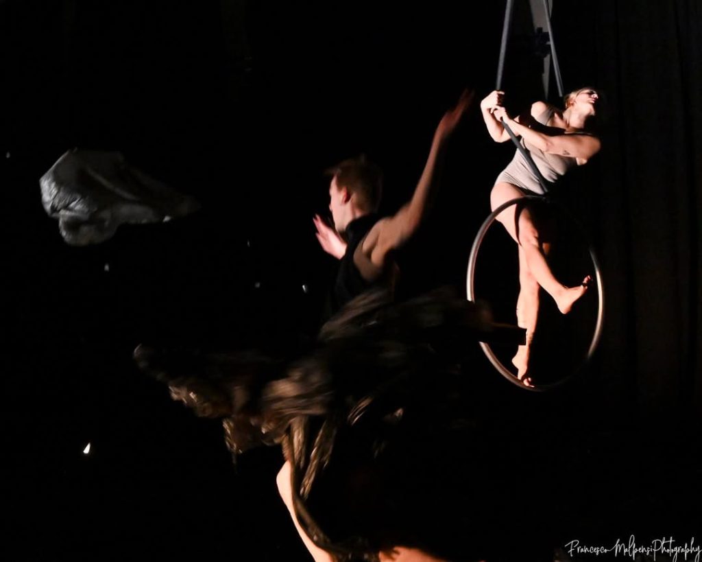2 artistes dansent. La première debout et le deuxième en dans un cerceaux en hauteur. Il est d’avantage éclairé.