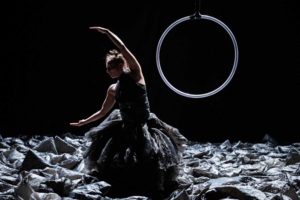 Habillée d’une de sacs plastiques une danseuse ouvre ses bras en demi-lune. Derrière elle se trouve un cerceau suspendu.