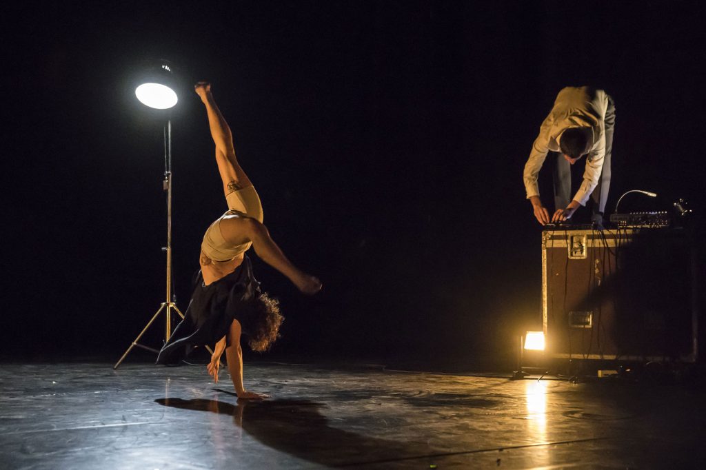 Deux circassiens, l’une en roue puis équilibre sur les mains à l’avant-scène, l’autre perché sur une caisse, éclairés par une lumière blanche et un contre chaud.