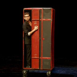 Un homme, sur scène, sort d'un casier en métal.