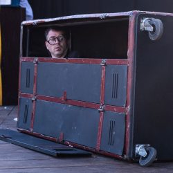 Un homme dans un casier en métal, vertical, laisse apercevoir la tête, lunettes penchées.