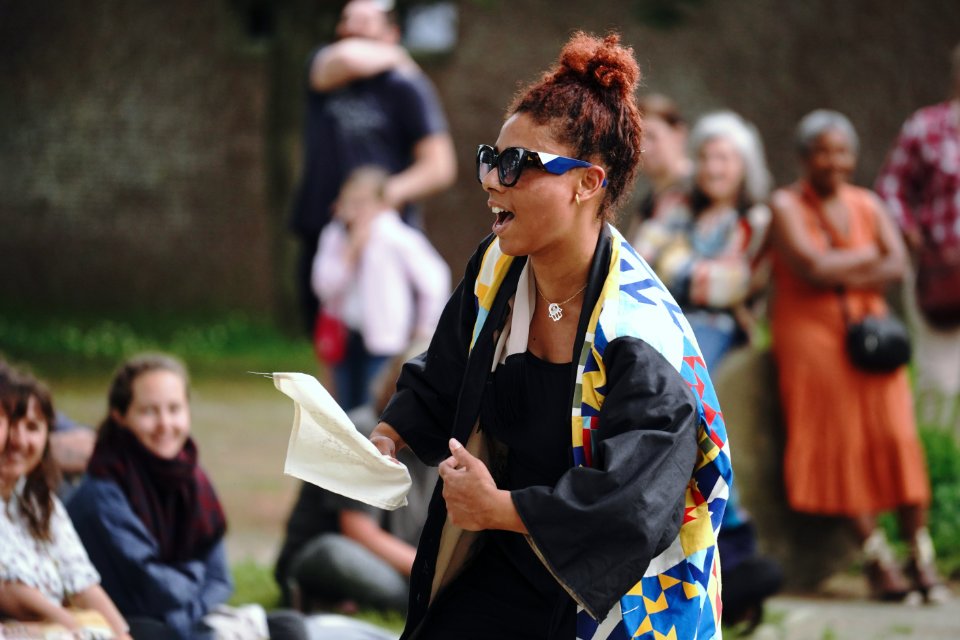 Une femme habillée en noir, veste multicolore et lunettes de soleil, tient un tissu. Elle est entourée du public.
