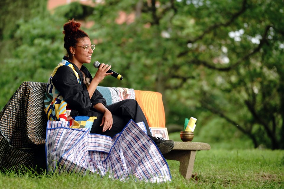 Une femme assise sur un banc, habillée en noir et veste patchwork multicolore. Elle parle au micro et a un grand sac à côté d'elle.