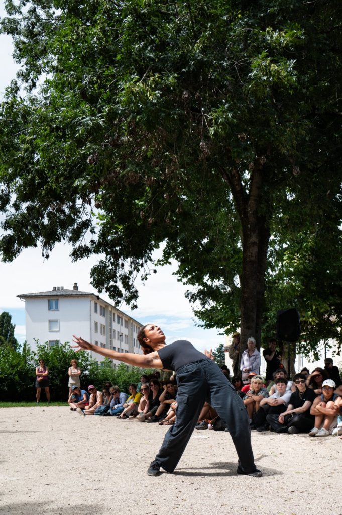 Une danseuse debout, tenue noire, cambrée et bassin en avant est entourée du public.