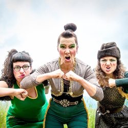 Dans un champ, 3 femmes habillées et maquillées en vert ont les points serrés et le visage crispé.