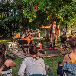 Dans un jardin, 2 hommes sont sur scène devant des spectateurs. L'un joue de la guitare et l'autre des claquettes