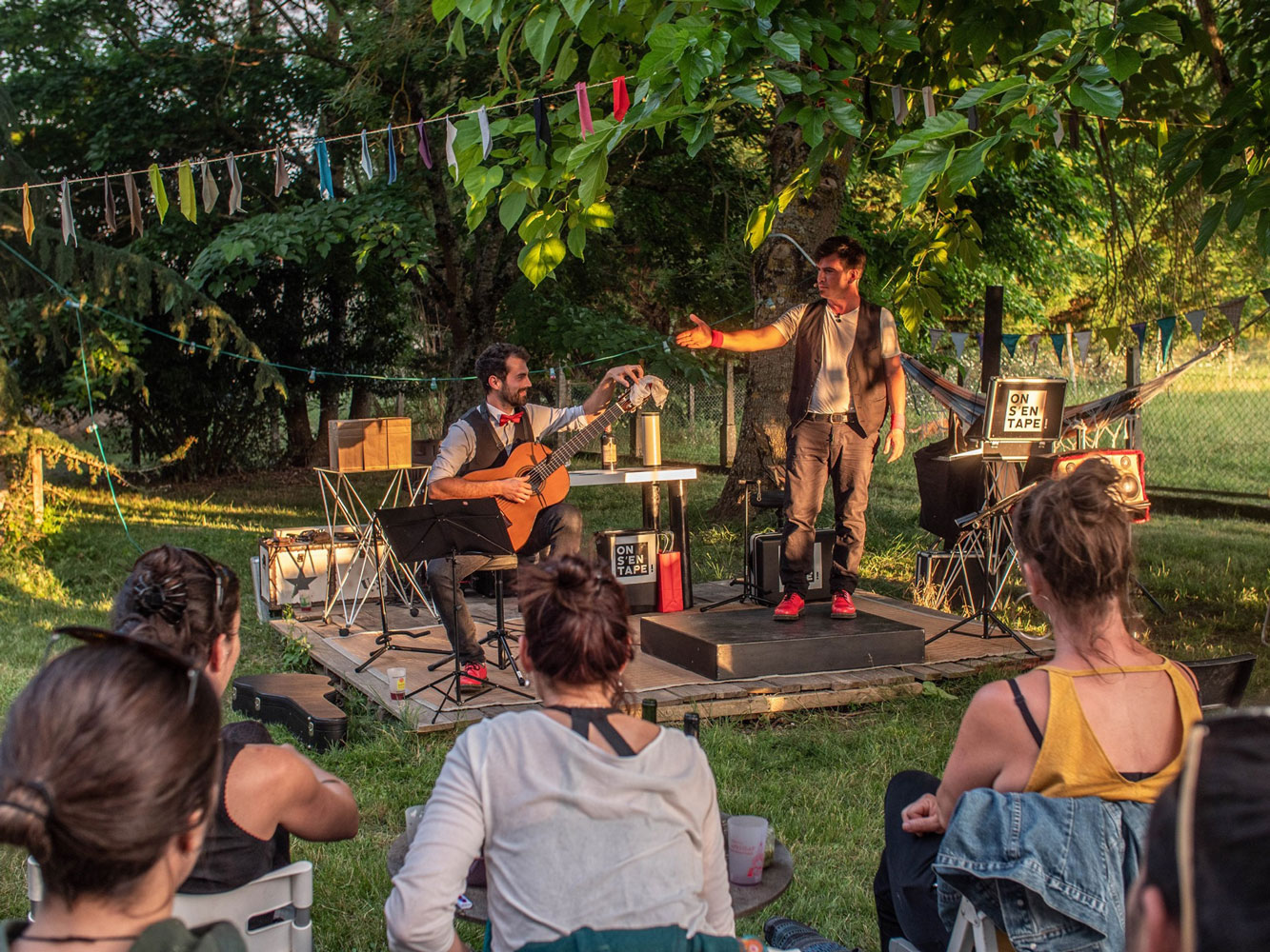 Dans un jardin, 2 hommes sont sur scène devant des spectateurs. L'un joue de la guitare et l'autre des claquettes