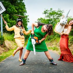 3 femmes en robes et tailleurs, sur une route de campagne tiennent un panneau, une tronçonneuse et une tête de licorne.
