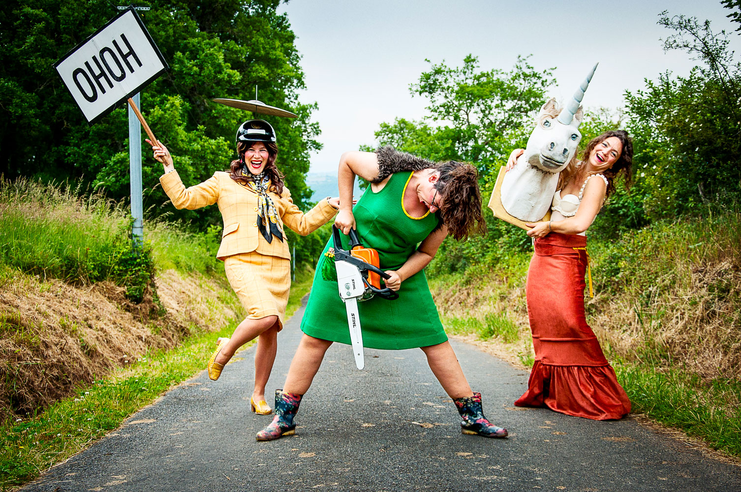 3 femmes en robes et tailleurs, sur une route de campagne tiennent un panneau, une tronçonneuse et une tête de licorne.
