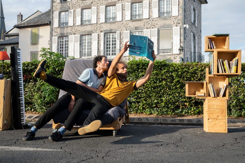 2 hommes lisent un livre en équilibre assis sur un banc en extérieur. Une petite bibliothèque est à leurs côtés.