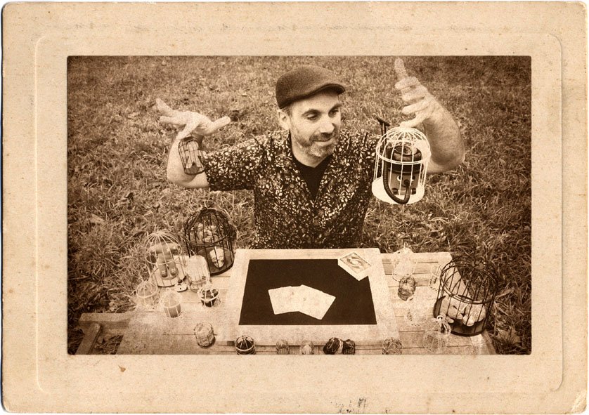 Tableau en sépia. Plusieurs cages à oiseaux sont posées sur la table ainsi que les cartes à jouer en tas et en éventail. Un homme tient une cage.