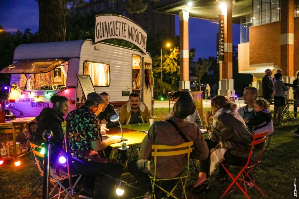 Plusieurs personnes attablées dans une guinguette assistent à un tour de magie.