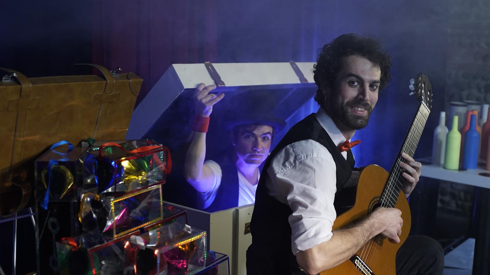 Sur scène, guitariste assis joue devant un homme qui sort d'une boite de magie.