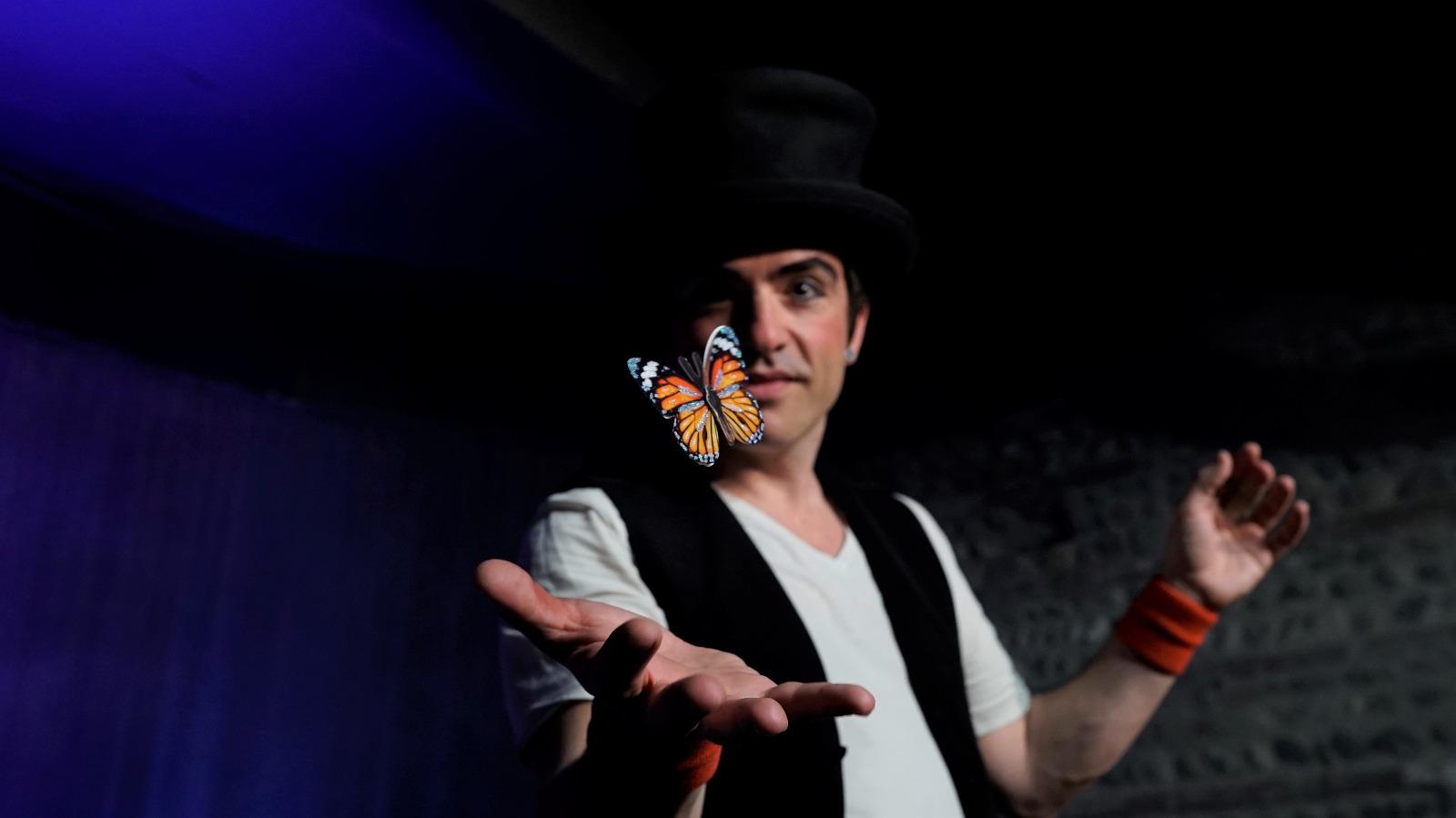 Un magicien tend la main et fait voler un papillon orange au-dessus de sa paume. Il le regarde.