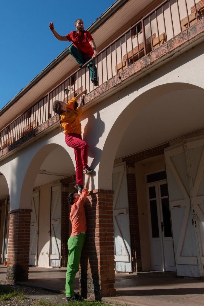 2 hommes se font la courte échelle et escaladent un mur d'immeuble. Le 3é me fait des acrobaties sur une balustrade.