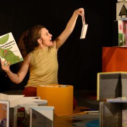 Une femme tient 2 livre de manière non conventionnelle. Elle est entourée d'une pyramide et d'un tourniquet de livre.