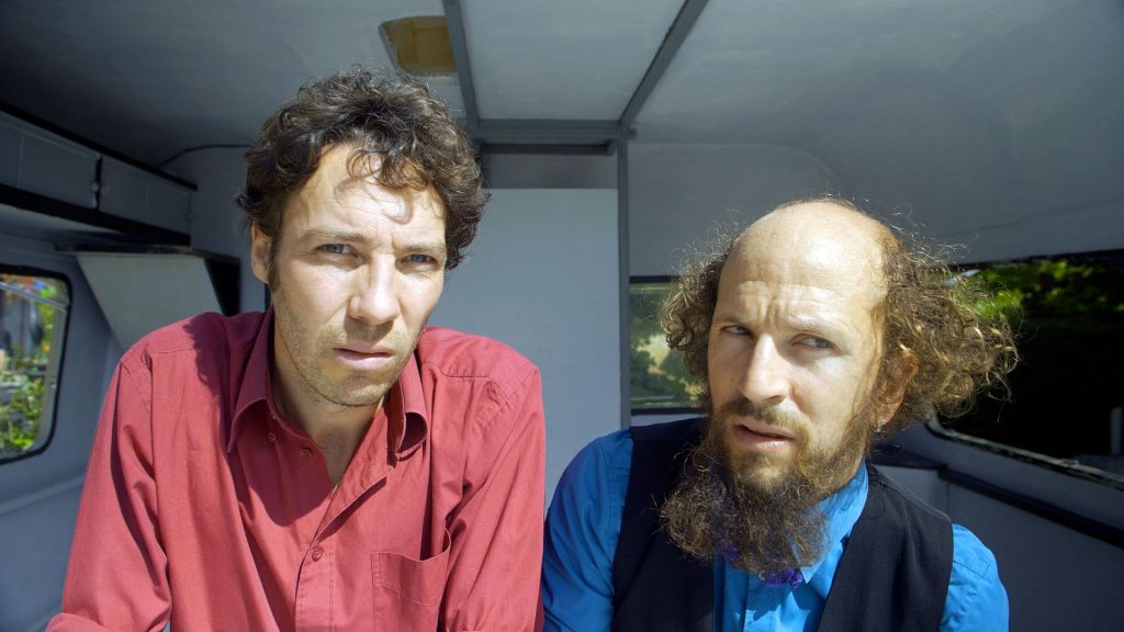 2 hommes dans une caravane. Le premier regarde droit devant et le deuxième a le regard un peu fuyant.