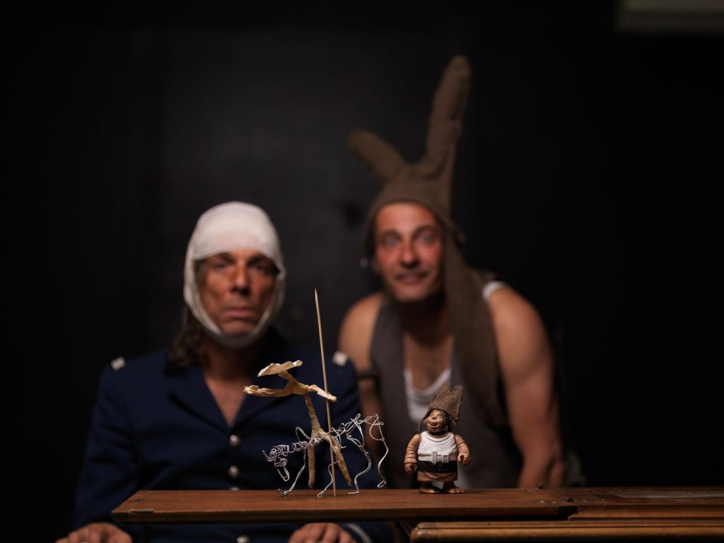 2 hommes, l'un avec une bande pensement sur la tête, l'autre avec un bonnet d'âne regarde une scénette avec des figurines en face d'eux.