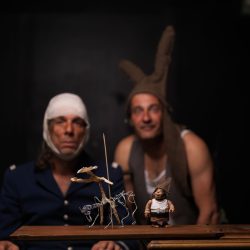 2 hommes, l'un avec une bande pensement sur la tête, l'autre avec un bonnet d'âne regarde une scénette avec des figurines en face d'eux.