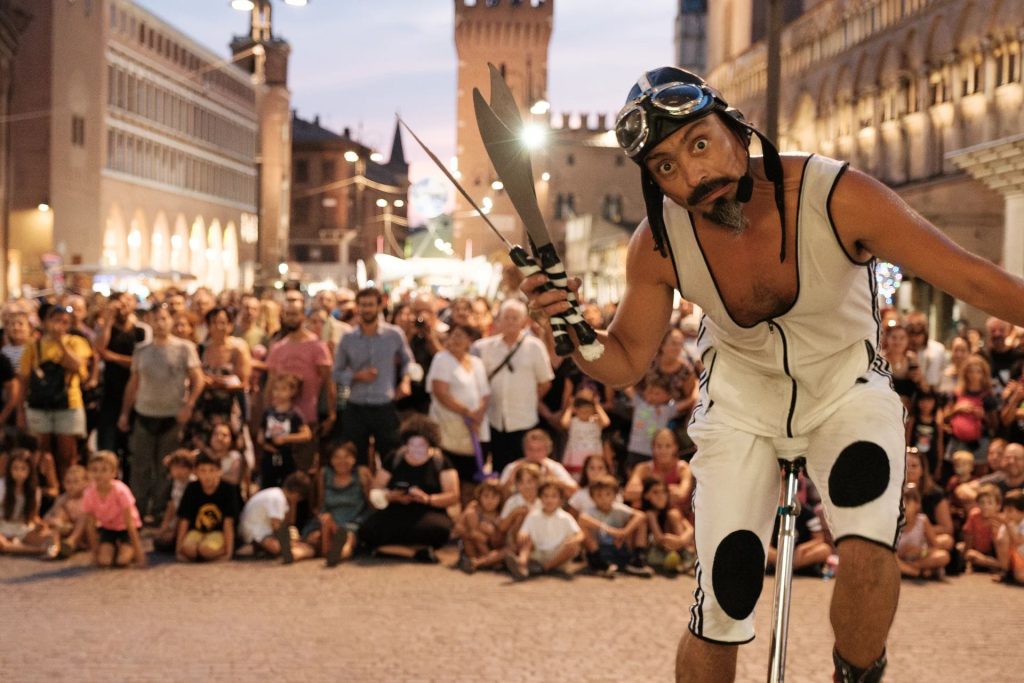 Un homme en cyclite blanc et noir, chapeau et lunettes de cycliste est au premier plan sur un monocycle, quilles de jongles à la main.