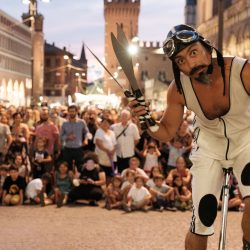 Un homme en cyclite blanc et noir, chapeau et lunettes de cycliste est au premier plan sur un monocycle, quilles de jongles à la main.