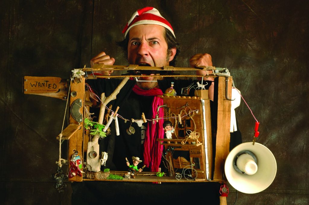 Un homme en bonnet bouche ouverte porte une petite valise dans lequel est construit une scénette en objets de récupération.