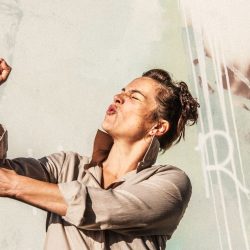 Une femme fait une grimace est a le poings levé. Derrière elle de la peinture et des graffitis.