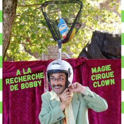 Un homme en costume et casque customisé avec une épuisette. Derrière lui un rideau de scène et un arbre.