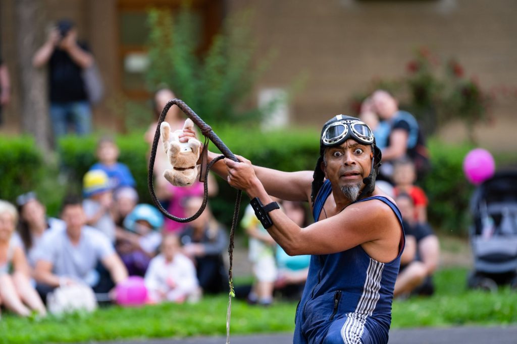 Un homme en lunettes et chapeau d'aviateur et débardeur bleu tient un lasso à la main dans lequel est une peluche. Le public le regarde.