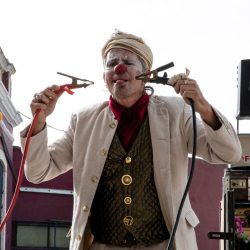 Clown maquillé et en beau costume tient des pinces crocodiles dans ses mains et fait une grimasse.