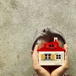 Un homme en bonnet se cache la tête avec une maison miniature qu'il tient dans ses mains.