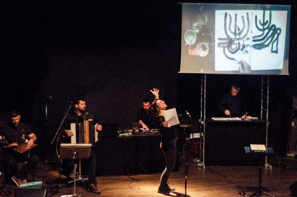 Des musiciens et une comédienne sont sur scène. Côté cours un dessinateur dessine et sont dessin est projeté sur un écran.