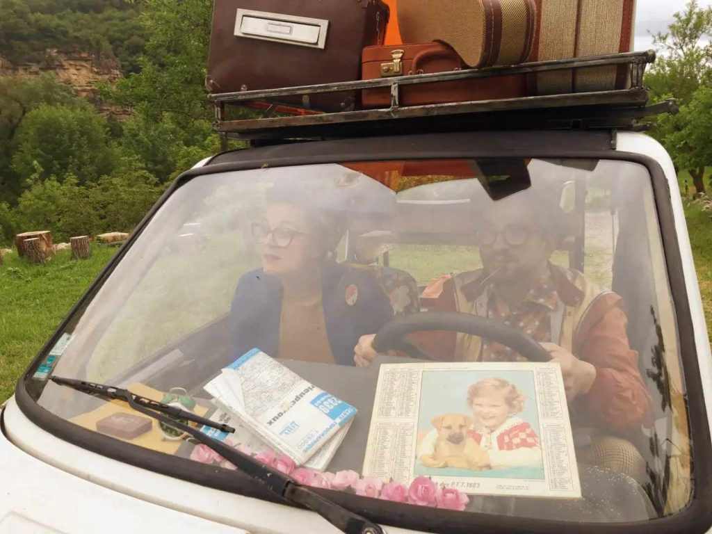Un couple dans les années 70 en voiture. On voit une carte routière et un calendrier sur le tableau de bord.