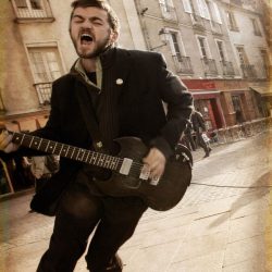 Un homme en costume chante dans la rue une guitare à la main.