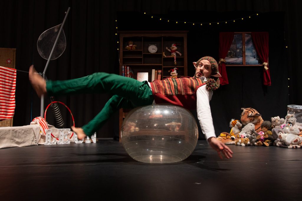 Sur scène un personnage de lutin tient l'équilibre sur un ballon de yoga.