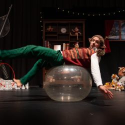 Sur scène un personnage de lutin tient l'équilibre sur un ballon de yoga.