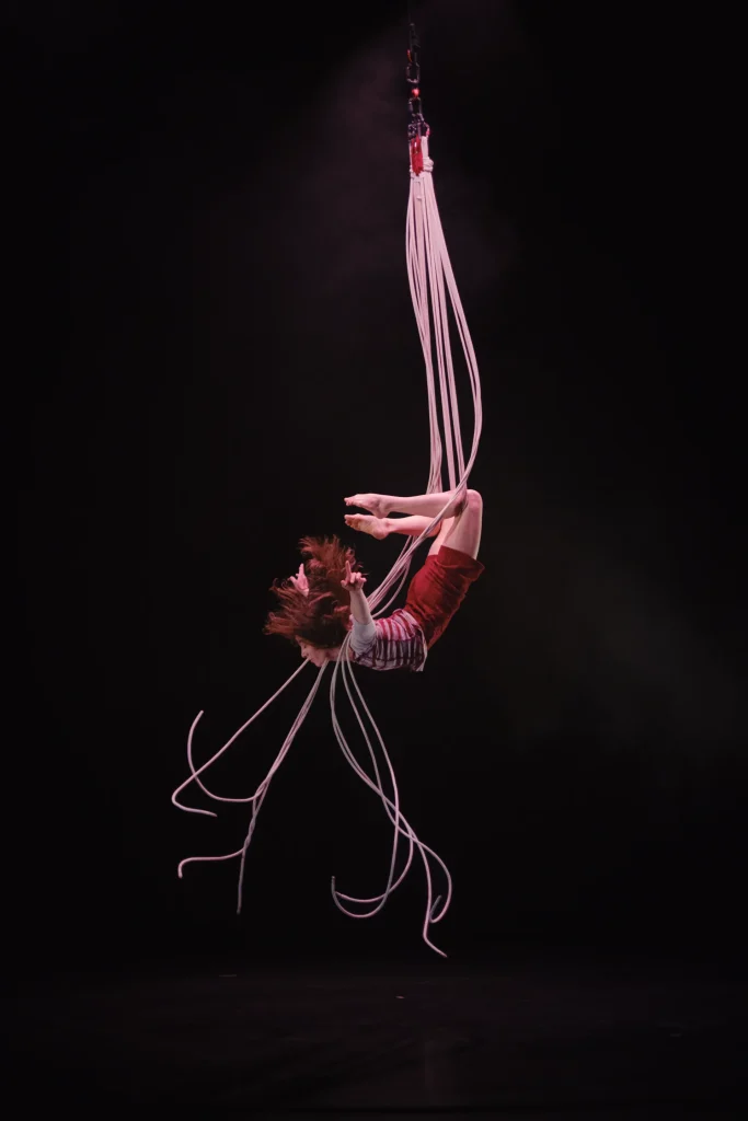 Une femme fait de la voltige sur scène. Elle est cambrée en hauteur, les lumières sont roses.