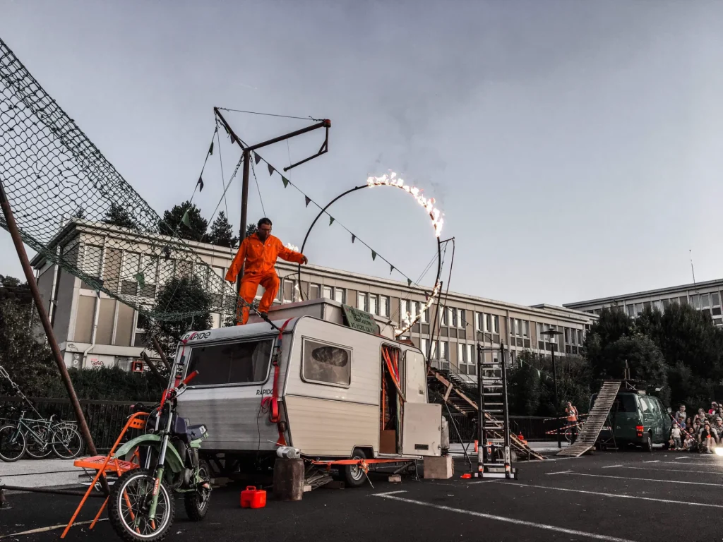 Un homme en tenue orange fait des cascades dans un parking, sur une caravane qui est entourée d'un ensemble d'agrégat de cirque et d'objets de tous les jours.