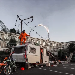 Un homme en tenue orange fait des cascades dans un parking, sur une caravane qui est entourée d'un ensemble d'agrégat de cirque et d'objets de tous les jours.