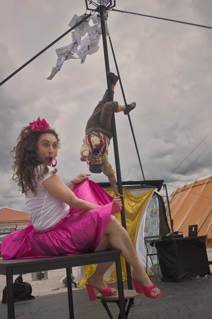 Des feuilles numérotées sont accrochées en haut d'un agrée de cirque. Une circassienne fait des acrobaties dans l'agrée, la tête en bas et une autre en robe est assise sur une table devant.