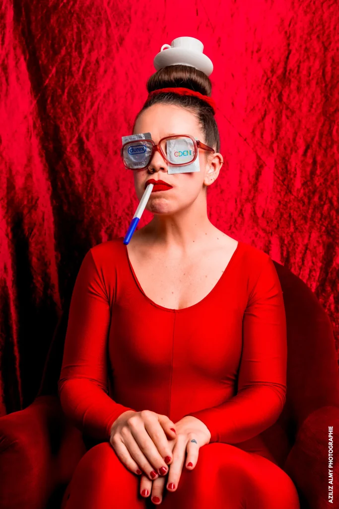 Une femme habillée en rouge avec vernis et chouchou rouge. Elle a des lunettes rouges dans lesquelles sont glissés des préservatifs et elle tient dans sa bouche un test de grossesse.