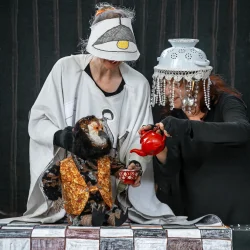 2 comédiennes en costume et chapeaux étranges (couscoussière avec des guirlandes de perles et casquette avec un dessin d'abat-jour) tiennent une marionnette chouette sur une table et servent un thé