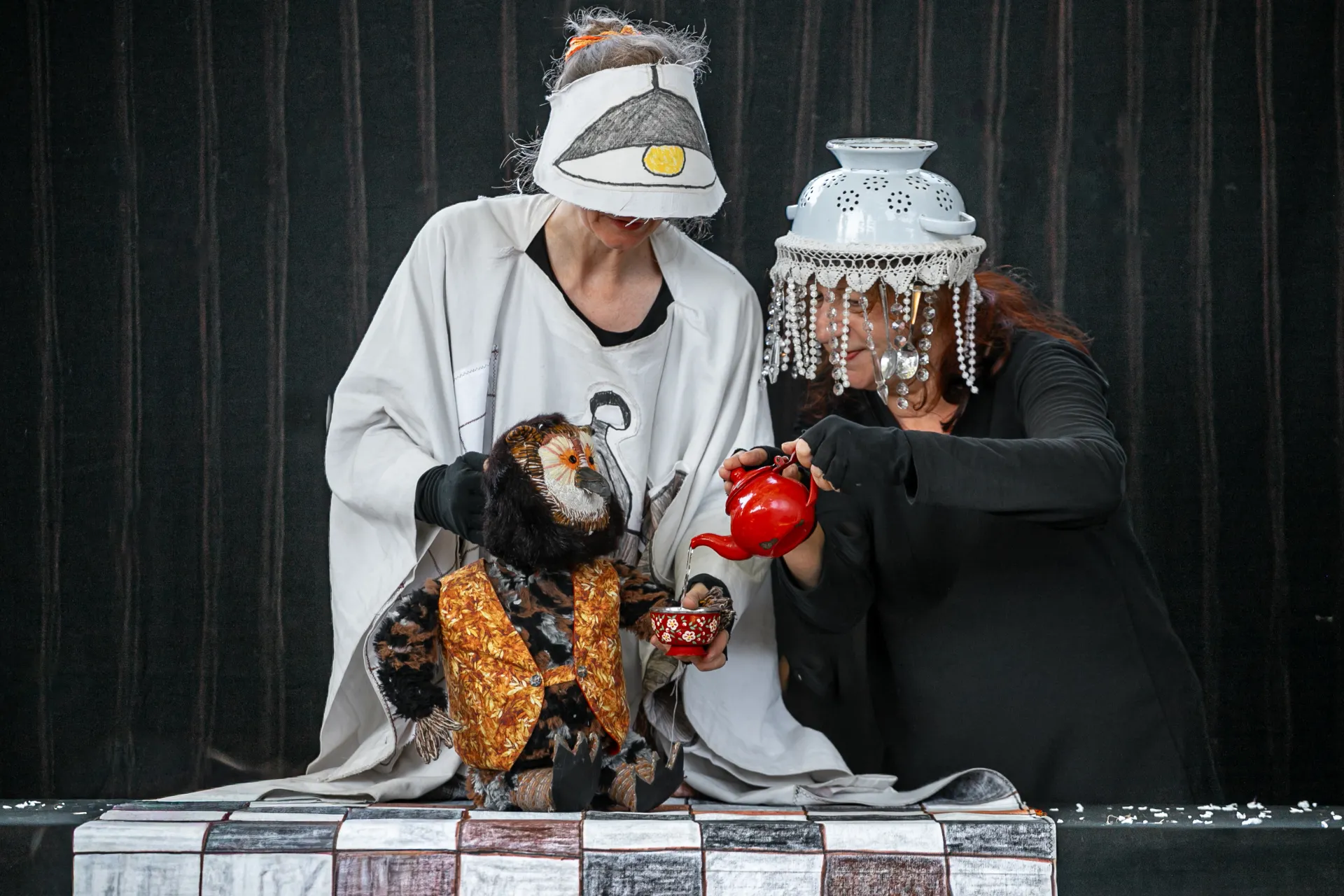 2 comédiennes en costume et chapeaux étranges (couscoussière avec des guirlandes de perles et casquette avec un dessin d'abat-jour) tiennent une marionnette chouette sur une table et servent un thé