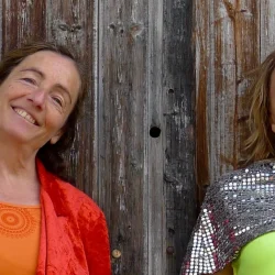Les 2 artistes de rouge virgule en costume colorés, et haut à facettes, pour une sourient. Elles sont adossées à un mur de bois.