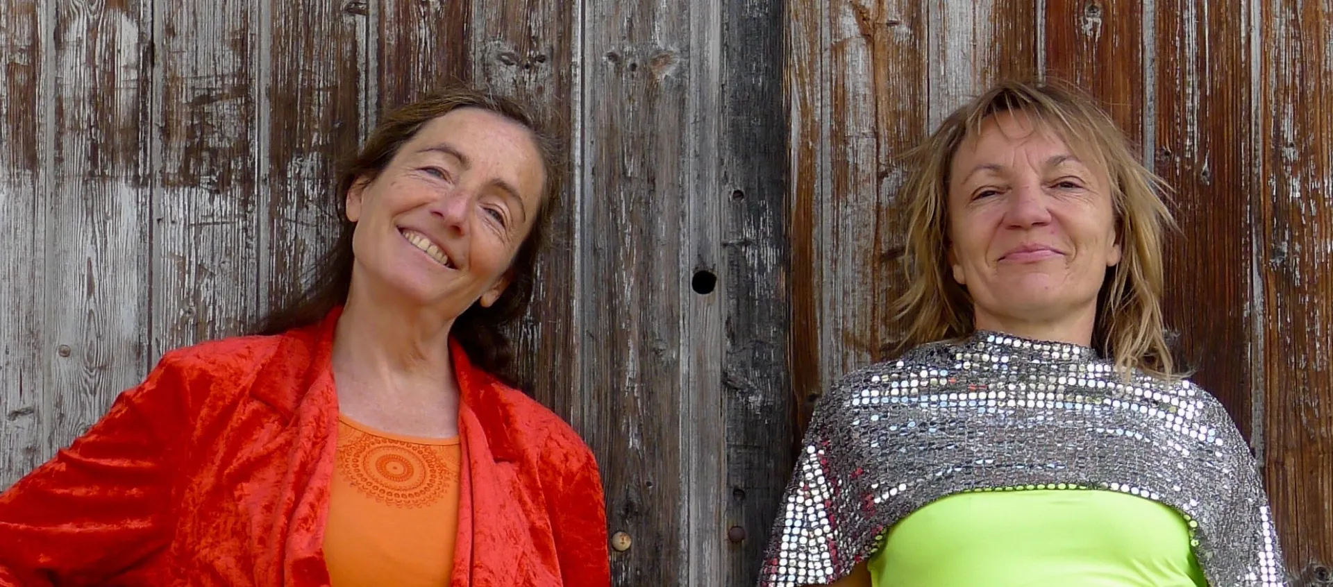 Les 2 artistes de rouge virgule en costume colorés, et haut à facettes, pour une sourient. Elles sont adossées à un mur de bois.
