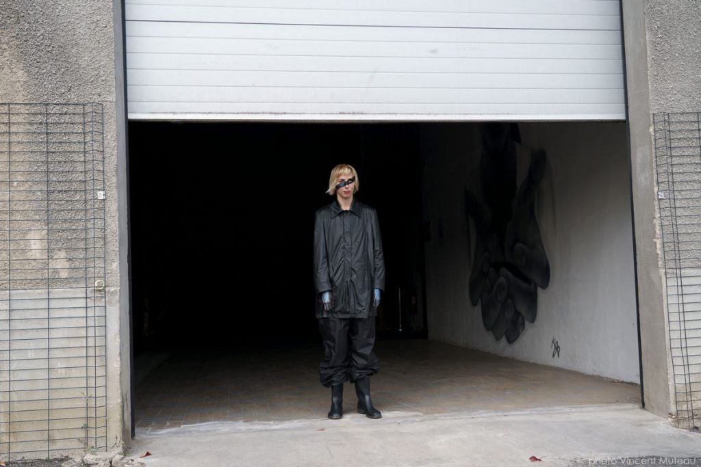 Une circassienne en noir sort d’une porte de garage entrouverte, maquillage bandeau, dans un décor industriel bétonné avec graffiti.