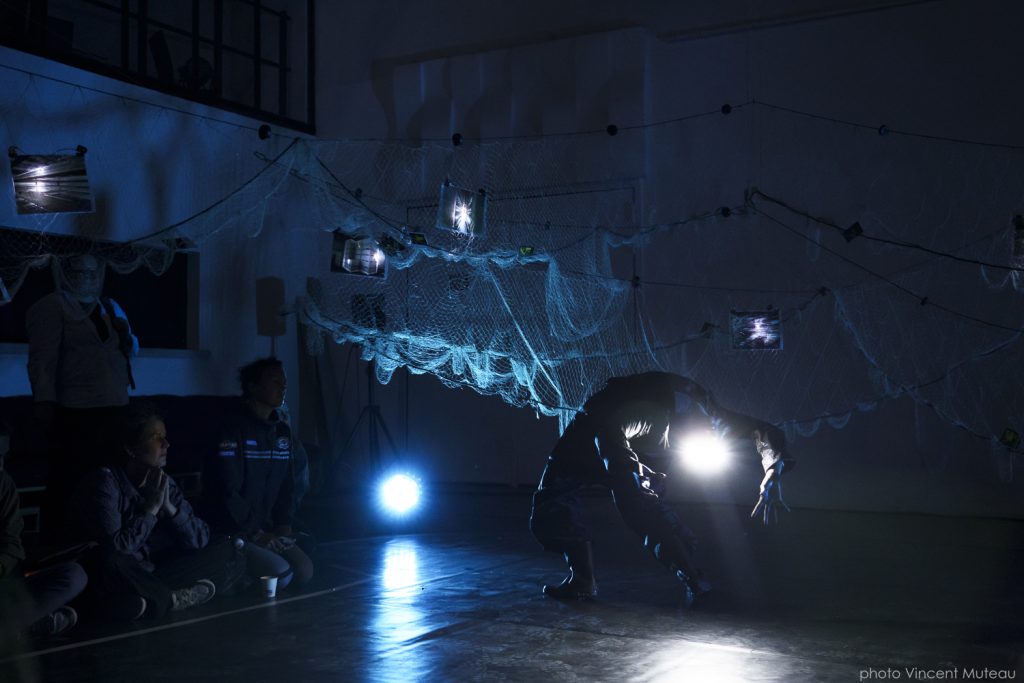 Dans une pièce sombre, une douce lumière bleue souligne des filets de pêche accrochés au mur. Une circassienne fait le pont.
