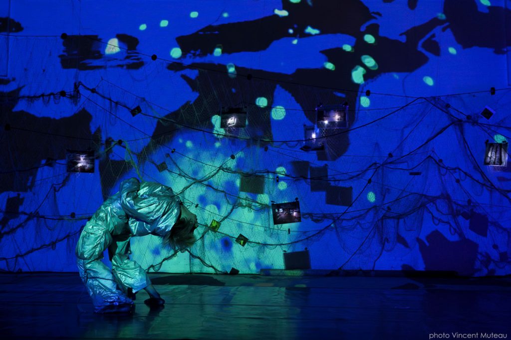 Une contorsionniste se cambre. Derrière elle, un mur remplit de filets de pêche et de photos éclairé en bleu.