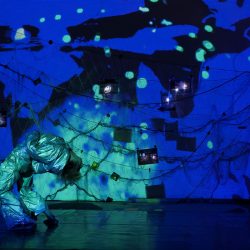 Une contorsionniste se cambre. Derrière elle, un mur remplit de filets de pêche et de photos éclairé en bleu.